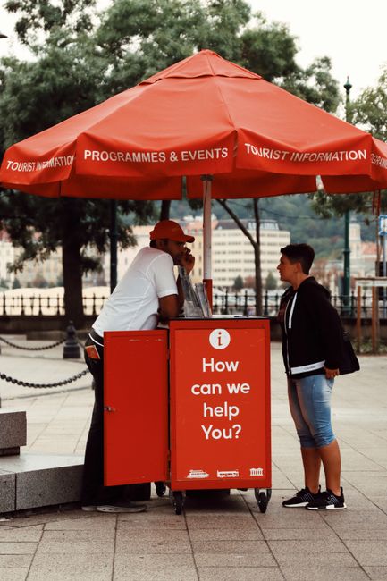 A street scene featuring a bright red tourist information kiosk with a matching large umbrella providing shade. The kiosk displays the message 'How can we help you?' in white text, and beneath it is a