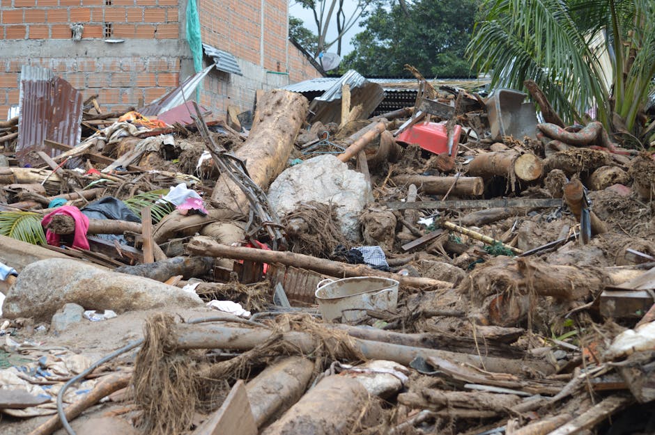 A garden scene following storm damage showing a chaotic mix of broken tree branches, uprooted logs, scattered debris, and overturned garden tools. The soil appears disturbed and muddy, with some brick